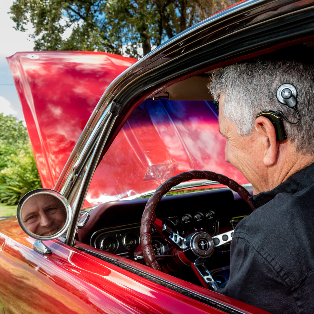 Rodney smiling and sitting in the driver's seat of his car listening to the engine with his cochlear implant visible behind his ear