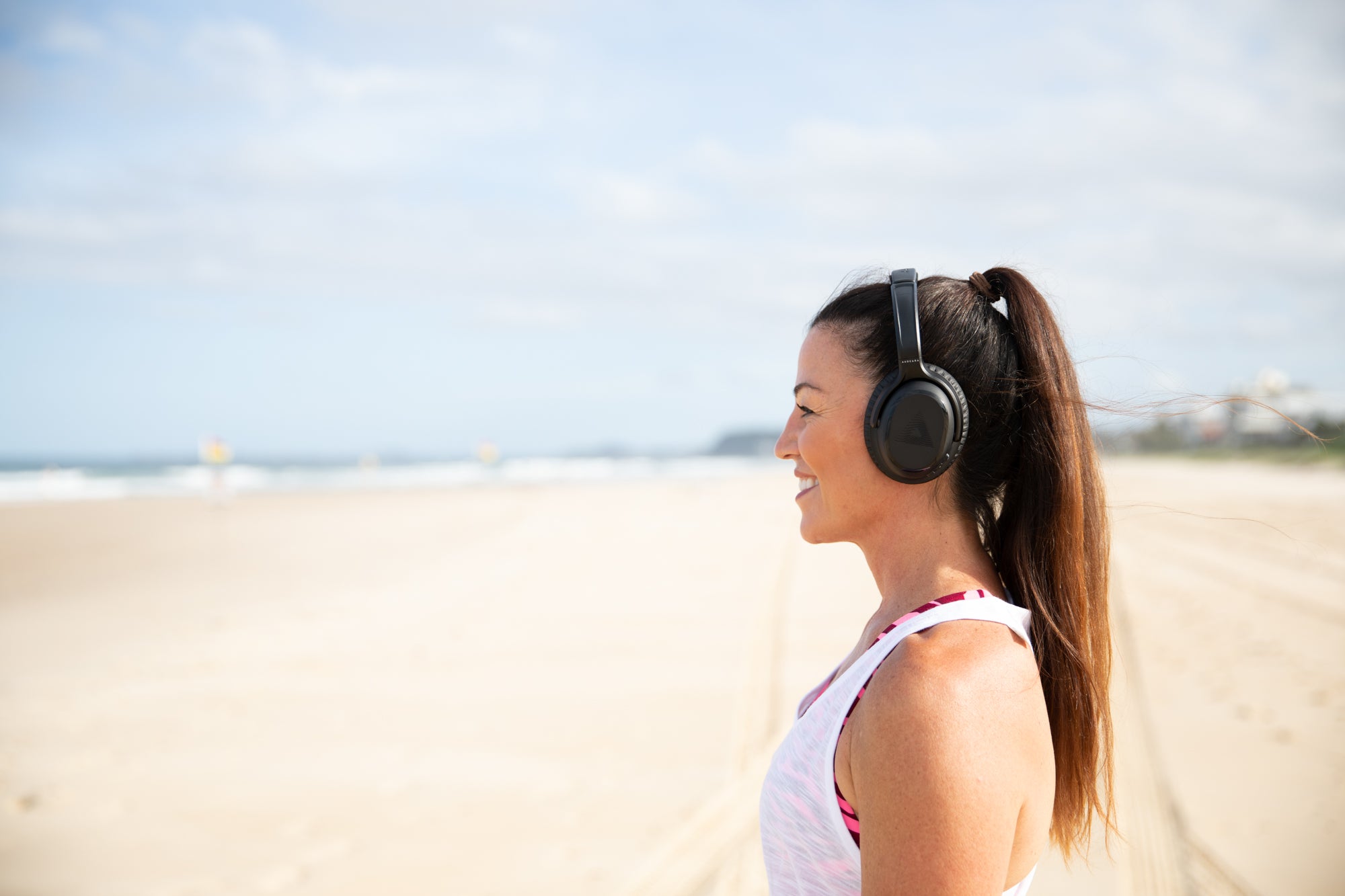 A woman smiling and wearing her Audeara headphones standing on the beach and looking out at the ocean