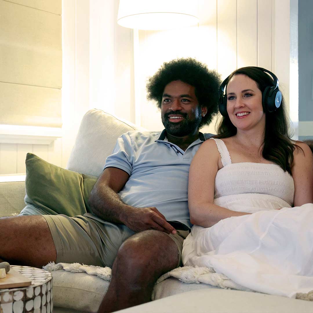 A woman wearing Audeara headphones and a man both sitting on the couch watching TV together