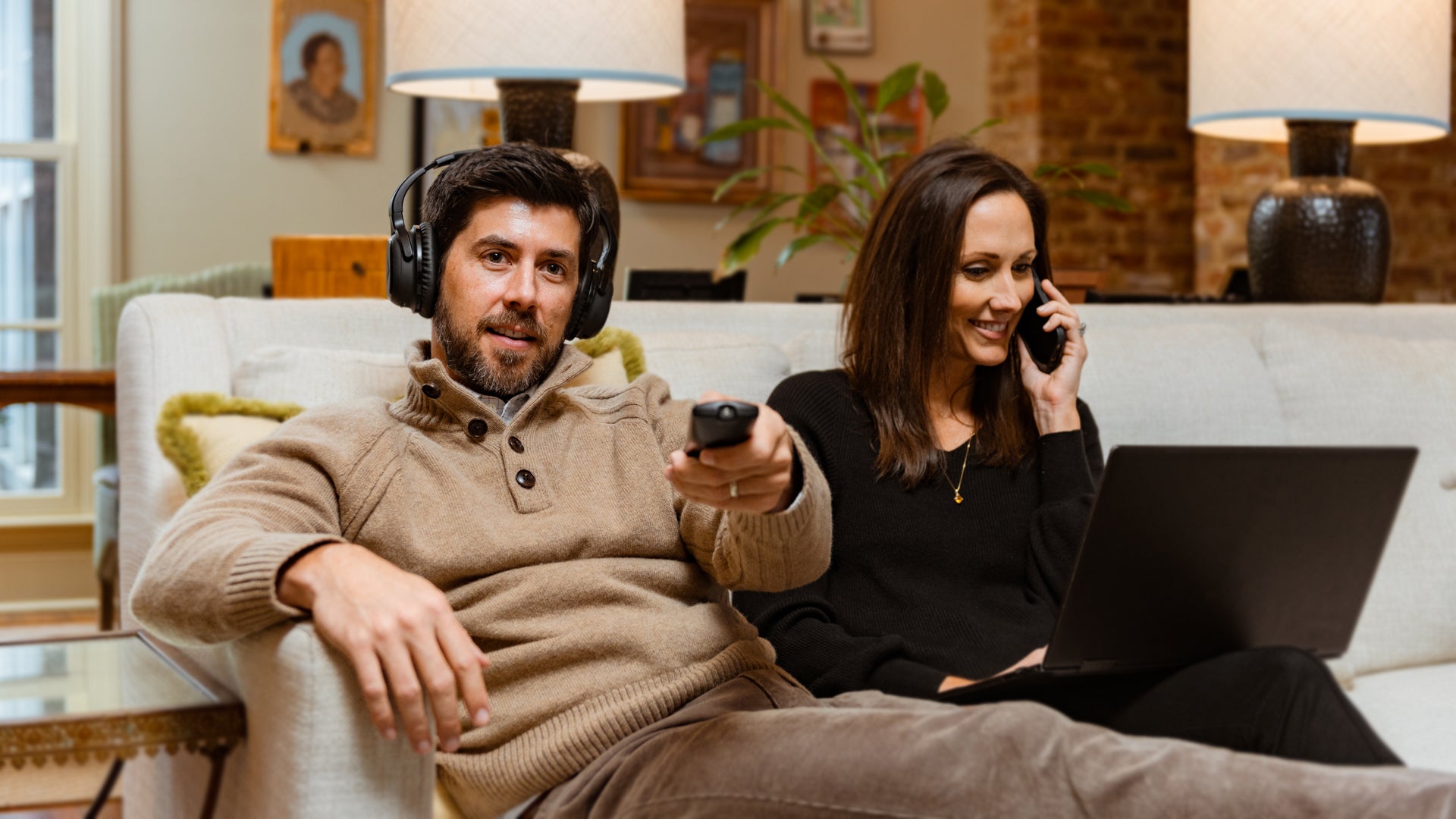 A man wearing Audeara headphones watches the TV while sitting next to his wife on the couch. The wife is on talking on the phone with a laptop in her lap.